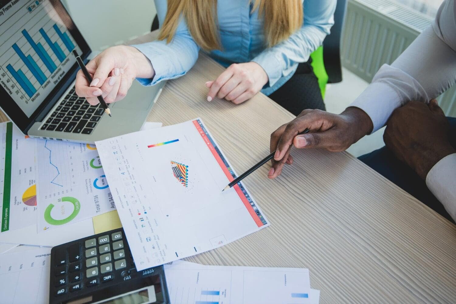 Pessoas em mesa com notebook, calculadora e papéis com relatórios