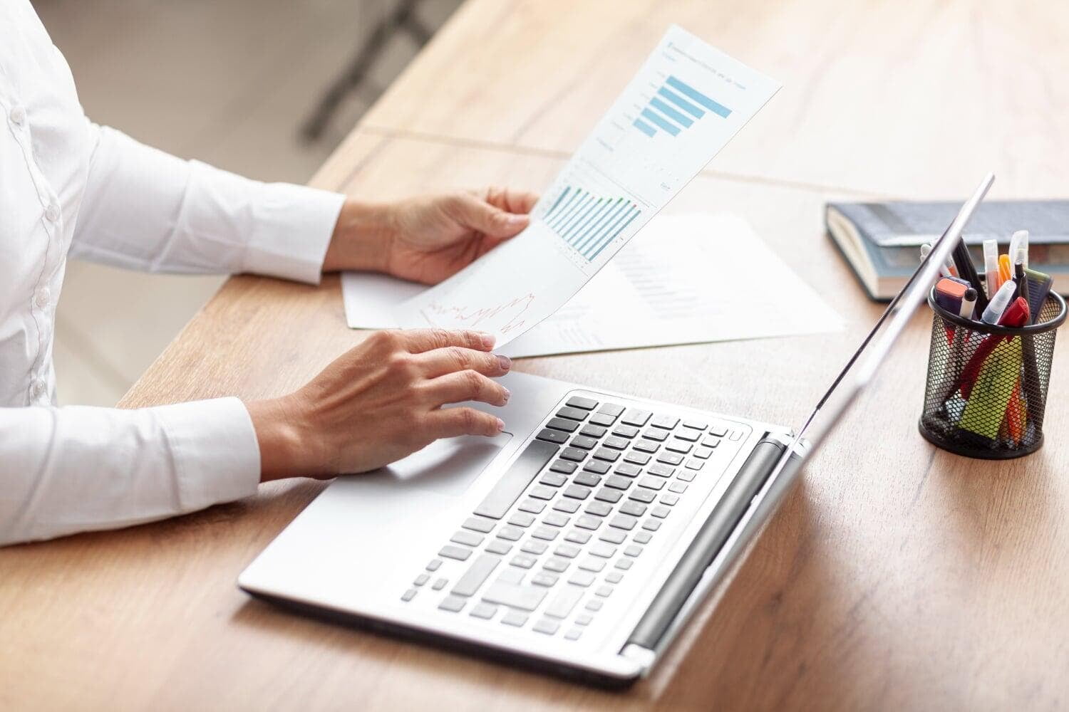 Pessoa em uma mesa com notebook segurando papel com relatórios e dados