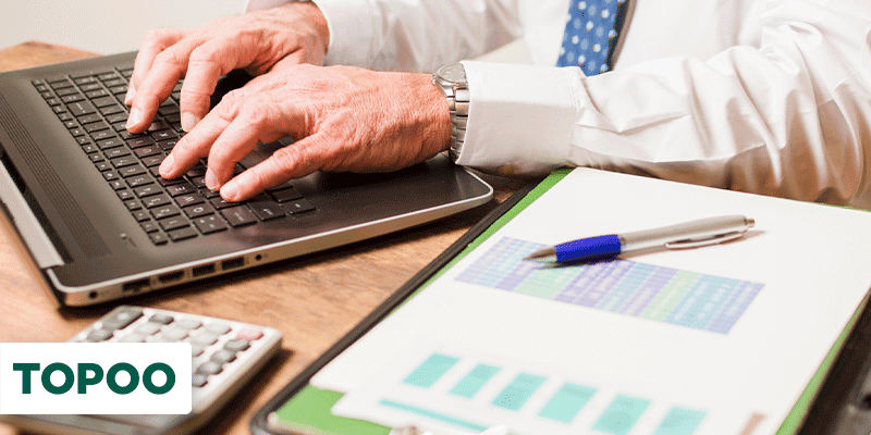 Homem sentado mexendo no notebook com papéis e uma calculadora na mesa