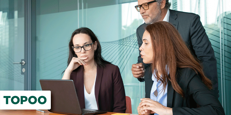 Um homem em pé e duas mulheres sentadas em uma mesa, todos olhando para um notebook