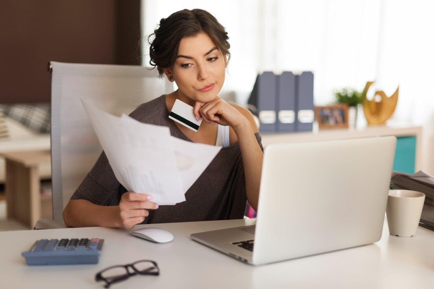 Mulher segurando cartão de crédito e papéis enquanto olha para o notebook