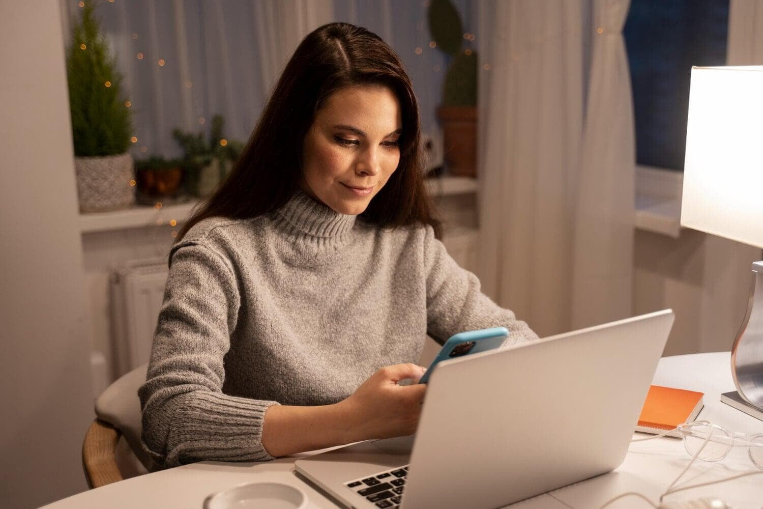 Mulher sorrindo enquanto olha um celular sentada em frente a um notebook